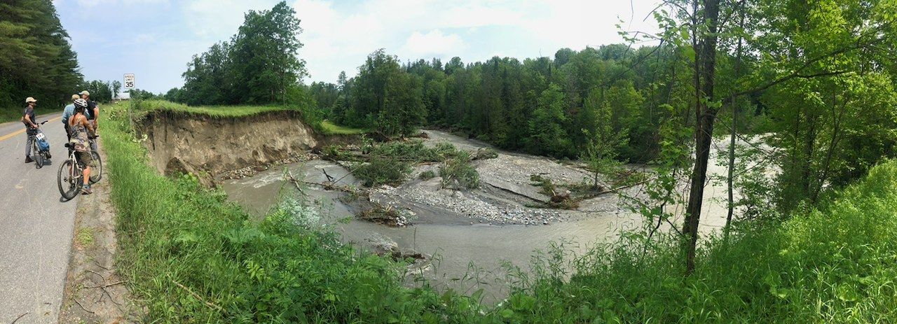 A wide view of a Vermont river that sheared off the road bank and cut a new gravel channel, with two people and a bike on the road beside a speed limit sign.