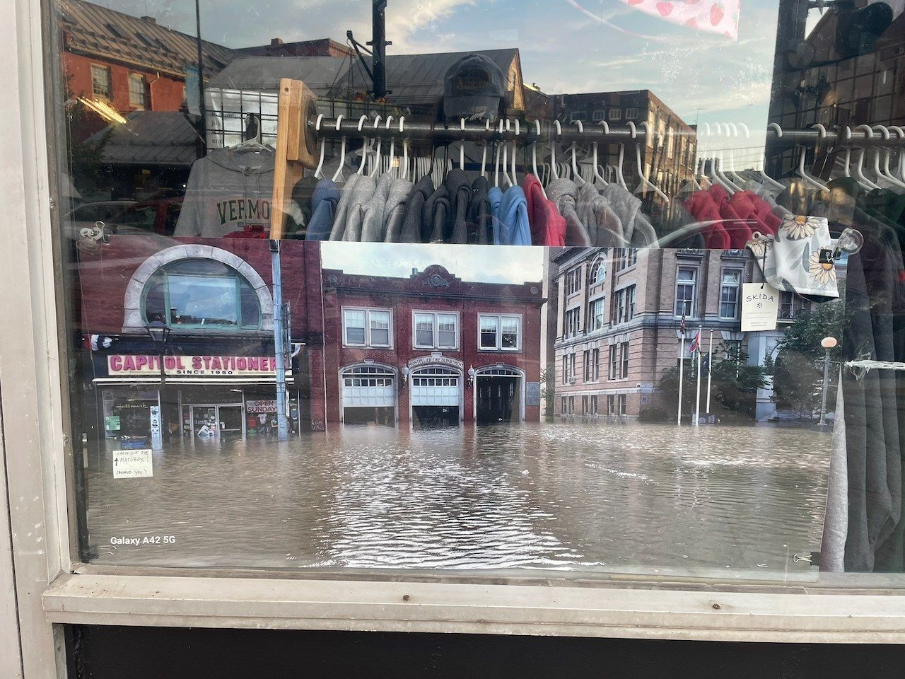 A print of downtown Montpelier under brown floodwater, displayed in a shop window with the present-day street reflected in the glass.